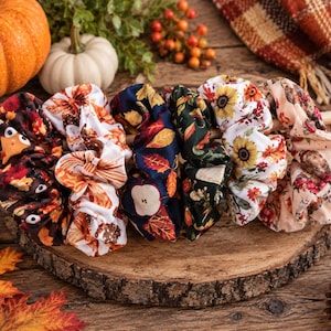 May include: A set of six fabric hair scrunchies featuring various autumn-themed patterns, including pumpkins, sunflowers, apples, and fall leaves in shades of orange, brown, white, and dark green, arranged in a row on a wooden dowel.
