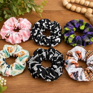 May include: Six fabric hair scrunchies arranged in two rows. The top row features a pink ghost print, a black cat and moon print, and a purple and green plaid pattern. The bottom row shows a colorful monster print, a white skeleton print, and a pumpkin print.