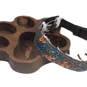 Peut inclure: Un plateau en bois brun foncé en forme d'empreinte de patte avec plusieurs compartiments, accompagné d'un collier pour animal avec un tissu bleu sarcelle foncé à motif floral orange et blanc, une sangle noire et des boucles en métal argenté.