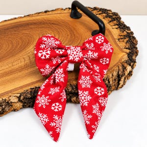 May include: A red fabric bow featuring a repeating white snowflake pattern. The bow has two long, pointed tails extending downwards and a central knot. The fabric is smooth and the snowflake designs vary in size and shape across the surface.
