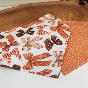 May include: A triangular pet bandana featuring a white background with a repeating pattern of brown and orange bows in various styles, including plaid, polka dot, and floral prints, paired with a solid orange side featuring white polka dots.