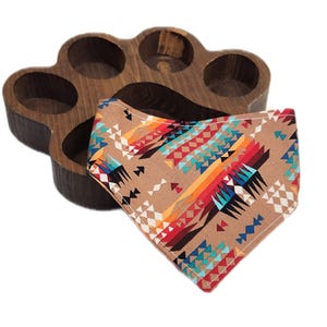 May include: A dark brown wooden tray shaped like a paw print with five circular indentations, paired with a triangular fabric bandana featuring a tan background and a colorful geometric pattern in shades of red, orange, yellow, blue, and white.