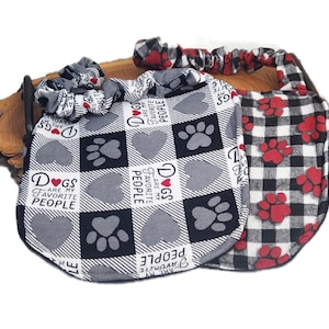 May include: Two fabric bibs with elastic neckbands. One bib features a black and white checkered pattern with gray hearts, paw prints, and the text "DOGS ARE MY FAVORITE PEOPLE". The other bib has a black and white buffalo plaid with red paw prints.