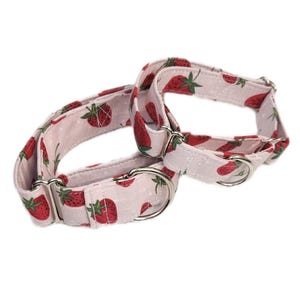 May include: Two dog collars featuring a strawberry print on a pale pink fabric. The collars have silver metal clasps and are designed for pets. The collars are made of fabric.