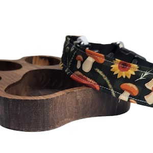 May include: A dark brown wooden paw-shaped bowl with four circular indentations positioned next to a black fabric pet collar featuring a repeating pattern of orange mushrooms, yellow sunflowers, and green foliage with a black plastic buckle.