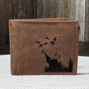 May include: Brown leather wallet with a stitched border. The wallet features a black silhouette of a hunter aiming a rifle at flying birds over tall grass. The wallet is rectangular and has a bi-fold design.