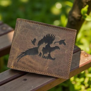 May include: Brown leather wallet with a detailed graphic of a hunter, birds, and a dog. The wallet has a stitched border and a weathered appearance, suggesting a vintage or rustic style. The design is etched into the leather.