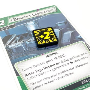 May include: A green and white card game card with a green background and a white border. The card features a picture of a laboratory with a character in the background. The card is titled "Banner's Laboratory" and has a number "2" in the top left corner. The card also has a black and yellow token with a "2" on it.