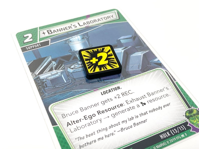 May include: A green and white card game card with a green background and a white border. The card features a picture of a laboratory with a character in the background. The card is titled "Banner's Laboratory" and has a number "2" in the top left corner. The card also has a black and yellow token with a "2" on it.