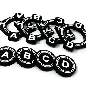 May include: Black and white game tokens featuring the letters A, B, C, and D. The tokens are in two styles: circular with a target design and angular with arrow designs. The letters are in a bold, sans-serif font.