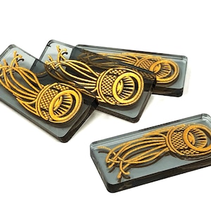 May include: Four rectangular glass tiles with a gold design of a stylised flower with a central circle. The tiles are stacked on top of each other.