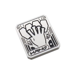 May include: A white enamel pin with a black outline of a hand with four fingers extended and a thumb tucked in. The hand is surrounded by a geometric design and the word "HAND" is printed below.