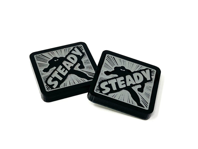 May include: Two black square-shaped acrylic tokens with a silver graphic of a person holding the word "STEADY" in a comic book style.
