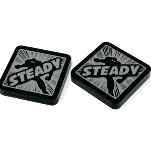 May include: Two black square-shaped tokens with a silver graphic of a superhero figure and the word "STEADY" printed on them.