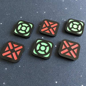 Może przedstawiać: Sześć kwadratowych żetonów do gry z czarnymi obramowaniami. Trzy żetony mają zielone koło z mniejszym czarnym kołem wewnątrz. Trzy żetony mają czerwony krzyż z czarnym obrysem.