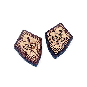 Könnte beinhalten: Zwei hölzerne Würfel in Diamantform. Die Würfel sind braun und haben eine hellbraune Holzmaserung. Die Würfel sind mit 1 und 2 nummeriert. Jeder Würfel hat eine kleine Illustration von zwei gekreuzten Äxten.