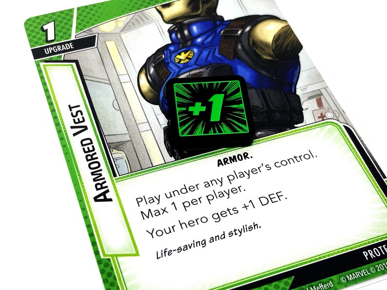 May include: A green and black card with the text "ARMORED VEST" and "UPGRADE" on it. The card has a green and black +1 symbol on it. The text on the card reads "ARMOR. Play under any player's control. Max 1 per player. Your hero gets +1 DEF. Life-saving and stylish."