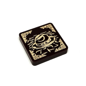 May include: A dark brown square tile with a gold design. The tile features a stylized image of a hat with a band and a wave pattern. The text "TILL" is written in gold lettering.