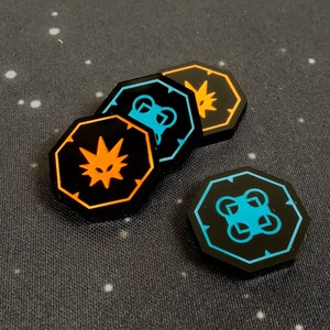 May include: Four black octagonal game tokens with different orange and blue symbols. The symbols include a creature with a star on its head, a creature with four eyes, a creature with a star on its head, and a four-square design.