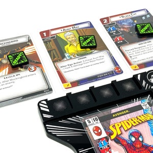 Puede incluir: Primer plano de un tablero de juego con cartas y dados. El tablero de juego es negro con rayas blancas y una portada de c&oacute;mic de Spiderman. Las cartas son del juego Marvel Champions y presentan personajes como Mockingbird, Aunt May y Black Cat. Los dados son verdes y tienen la palabra "READY!" en ellos.