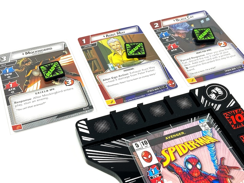 Puede incluir: Primer plano de un tablero de juego con cartas y dados. El tablero de juego es negro con rayas blancas y una portada de c&oacute;mic de Spiderman. Las cartas son del juego Marvel Champions y presentan personajes como Mockingbird, Aunt May y Black Cat. Los dados son verdes y tienen la palabra "READY!" en ellos.