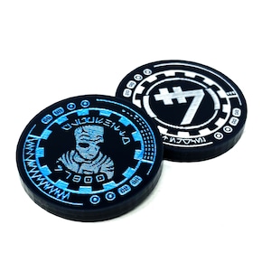 Könnte beinhalten: Zwei schwarze runde Token mit einem weißen und blauen Design. Ein Token zeigt die Nummer 7 in Silber auf weißem Hintergrund. Der andere Token zeigt eine Schwarzweißillustration einer Person auf blauem Hintergrund und den Text "7900".