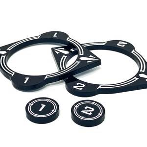 Könnte beinhalten: Zwei schwarze Plastik-Spielsteine mit weißen Zahlen und Pfeilen. Die Steine sind ringförmig mit einem spitzen Ende. Es gibt auch zwei kleinere schwarze Plastik-Token mit weißen Zahlen.