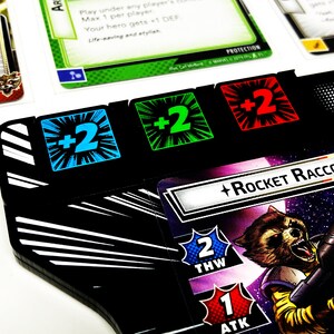 May include: A black and white game board with three counters showing +2. The counters are blue, green, and red. A card with the text "Rocket Raccoon" and a picture of Rocket Raccoon is in the background.