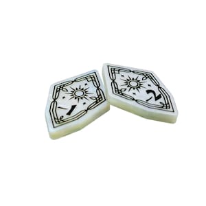 May include: Two white, diamond-shaped game pieces with black designs. One piece has the number "1" and the other has the number "2".