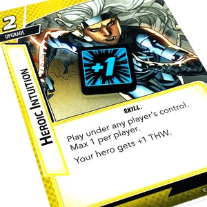 May include: A yellow card with a comic book character image and the text "HEROIC INTUITION" and "SKILL." The card has a blue and black +1 token on it. The text on the card reads "Play under any player's control. Max 1 per player. Your hero gets +1 THW."