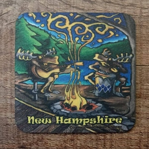 May include: Square coaster featuring a whimsical illustration of two moose playing music around a campfire. The background includes a lake, trees, and a starry sky. The text "New Hampshire" is displayed at the bottom.