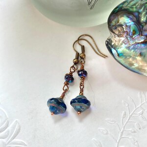 Puede incluir: Pendientes hechos a mano con cuentas facetadas azules y moradas y detalles de alambre de cobre. Los pendientes tienen una estética vintage con una combinación de cuentas azul oscuro e iridiscentes. Los ganchos para las orejas son de color bronce.