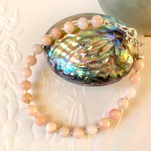 May include: A delicate bracelet featuring round, peach-colored beads strung together with silver-toned metal accents. The bracelet is displayed on a shimmering, iridescent abalone shell, creating a contrast of textures and colors. The bracelet is a piece of jewelry.