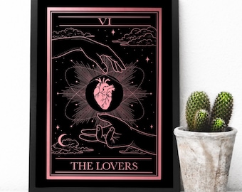Puede incluir: Una impresión enmarcada en negro de la carta del tarot de Los Amantes con un diseño en oro rosa. La carta presenta dos manos que se acercan a un corazón en un círculo. El texto "VI THE LOVERS" está en la parte inferior de la carta.