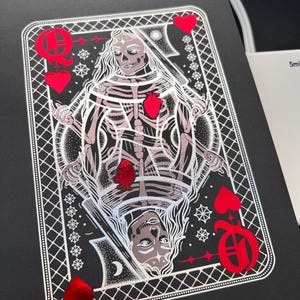 May include: A black card with a skeletal Queen of Hearts design. The illustration is silver and red, with the Queen holding a sword. The card is framed with a white, geometric border and red heart suit symbols.