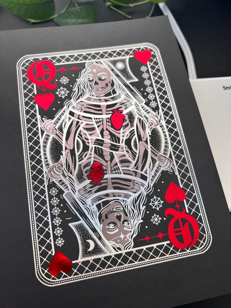 May include: A black card with a skeletal Queen of Hearts design. The illustration is silver and red, with the Queen holding a sword. The card is framed with a white, geometric border and red heart suit symbols.