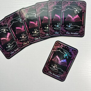 May include: A set of six holographic tarot cards with a black background and pink floral accents. The cards feature a hand holding an open book with a crescent moon design. The text on the cards reads "The Reader".