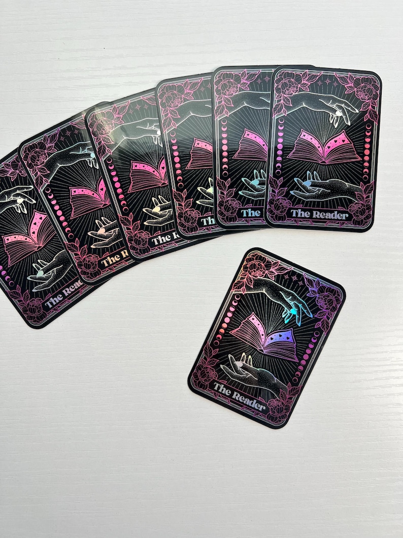 May include: A set of six holographic tarot cards with a black background and pink floral accents. The cards feature a hand holding an open book with a crescent moon design. The text on the cards reads "The Reader".