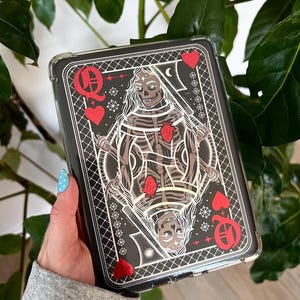 May include: Black phone case with a Queen of Hearts playing card design. The card illustration features a skeleton figure with a red heart, surrounded by red hearts and card suit symbols. The case has a clear, protective border.