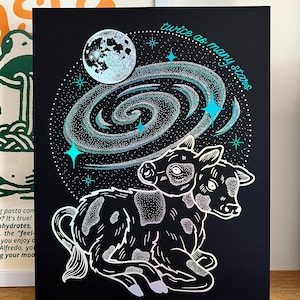 May include: Black art print with a two-headed cow illustration, a galaxy, and a moon. The teal text reads "twice as many stars". The cow is white with black spots, set against a black background. The artwork has a celestial theme.