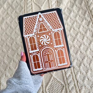 Puede incluir: Una funda transparente para teléfono con un diseño de casa de jengibre. La funda tiene un fondo oscuro con una ilustración detallada de una casa de jengibre en tonos marrones y blancos. El diseño incluye un tejado, ventanas y una puerta.