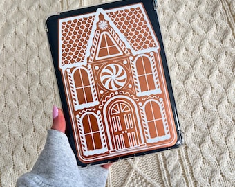 May include: A clear phone case featuring a gingerbread house design. The case has a dark background with a detailed gingerbread house illustration in shades of brown and white. The design includes a roof, windows, and a door.