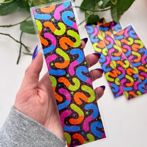 May include: A clear bookmark with a colorful design. The design features a black background with a grid pattern and vibrant, curved shapes in neon pink, orange, green, and blue, along with small star shapes. The bookmark is held in a hand.