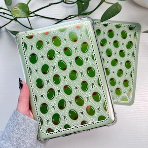 Puede incluir: Dos fundas para teléfono con un diseño de aceitunas verdes. Las fundas tienen un fondo verde claro con un patrón repetido de aceitunas verdes con centros rojos, conectadas por lazos y líneas blancas. Las fundas están en fundas protectoras transparentes.