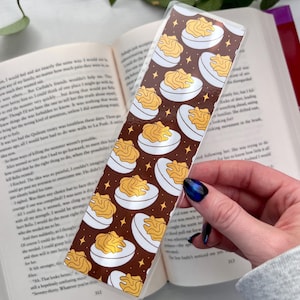 May include: A rectangular bookmark featuring a repeating pattern of deviled eggs on a dark brown background with gold stars. The bookmark is held over an open book, with the page number 312 visible.
