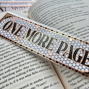 May include: A decorative bookmark with the text "ONE MORE PAGE" in black letters. The bookmark has a mosaic-like design with black and gray tiles, outlined with a gold border. The bookmark is placed on top of an open book.