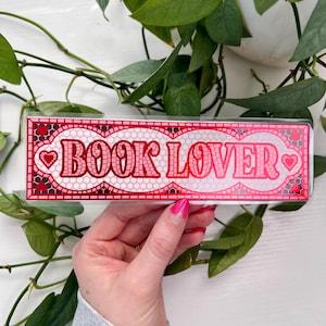 以下が含まれることがあります： 装飾的なフォントで「BOOK LOVER」という言葉が入った長方形のブックマーク。 ブックマークは、ハートのアクセントが付いたピンクと赤のモザイクデザインです。 背景には緑の葉の植物と白い表面が含まれています。