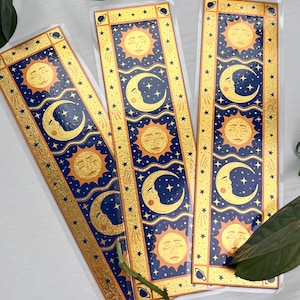 May include: Three celestial-themed bookmarks. Each bookmark features a blue background with gold accents, sun and moon illustrations, and star details. The bookmarks are rectangular and have a glossy finish.