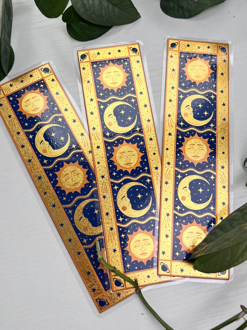 May include: Three celestial-themed bookmarks. Each bookmark features a blue background with gold accents, sun and moon illustrations, and star details. The bookmarks are rectangular and have a glossy finish.