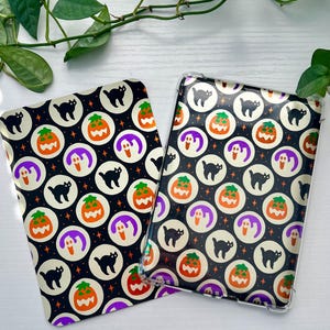May include: Two Halloween-themed phone cases with a black background and a repeating pattern of orange pumpkins, black cats, and purple ghosts. The cases are made of clear plastic and have a glossy finish.